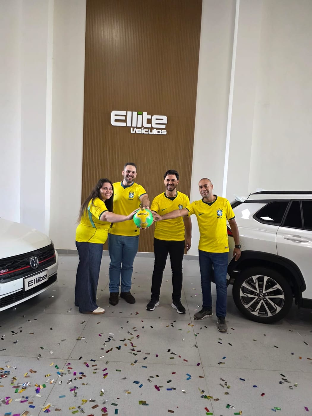 Quatro integrantes da equipe da Ellite Veículos de camisa da seleção brasileira, segurando uma bola no salão entre dois carros, com confete no chão