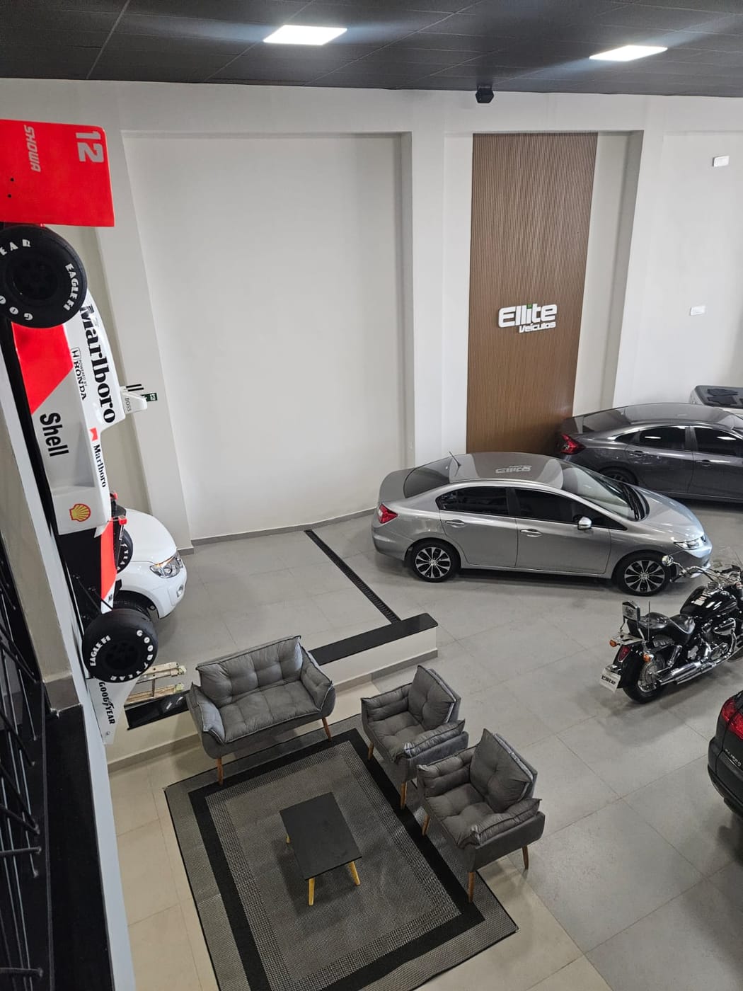 Salão de exposição visto de cima, do mezanino, com a réplica da McLaren à esquerda, um Honda Civic prata ao centro e uma Harley-Davidson à direita