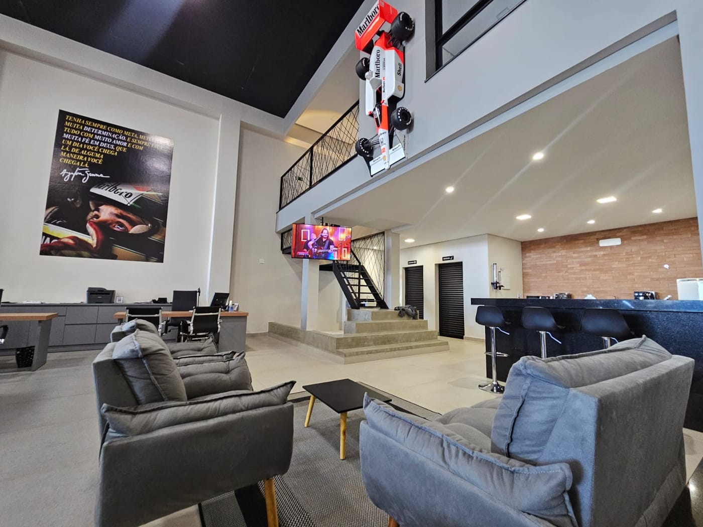 Lounge de espera com poltronas cinza e a réplica em tamanho real da McLaren MP4/4 de Ayrton Senna suspensa na borda do mezanino