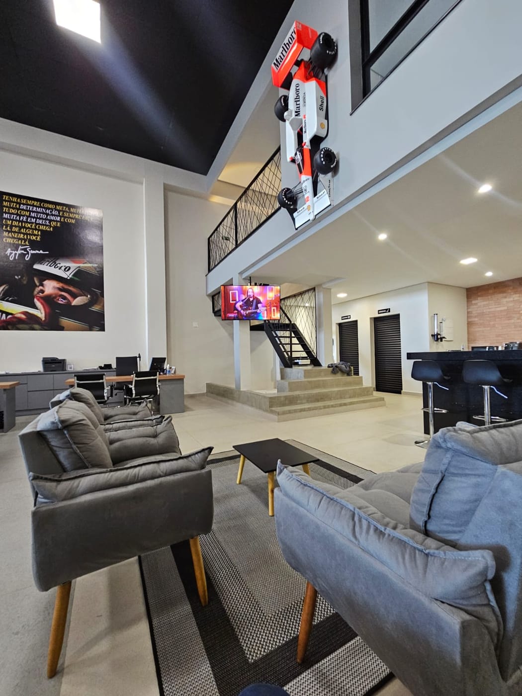 Sala de espera com o quadro de Ayrton Senna e sua frase sobre determinação na parede, a escada para o mezanino ao fundo e a réplica da McLaren suspensa acima